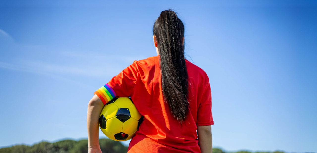 Foto van de achterkant van een persoon met een voetbal onder diens arm en een regenboogband onderaan de mouw