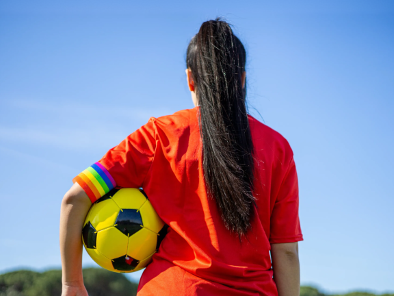 Foto van de achterkant van een persoon met een voetbal onder diens arm en een regenboogband onderaan de mouw